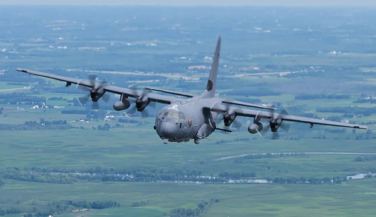 A large military plane flying over the top of a green field.